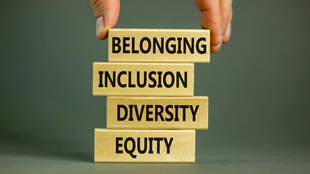 A hand placing a wooden building block onto a small stack that has the words 'Equity', 'Diversity', 'Inclusion' and 'Belonging'