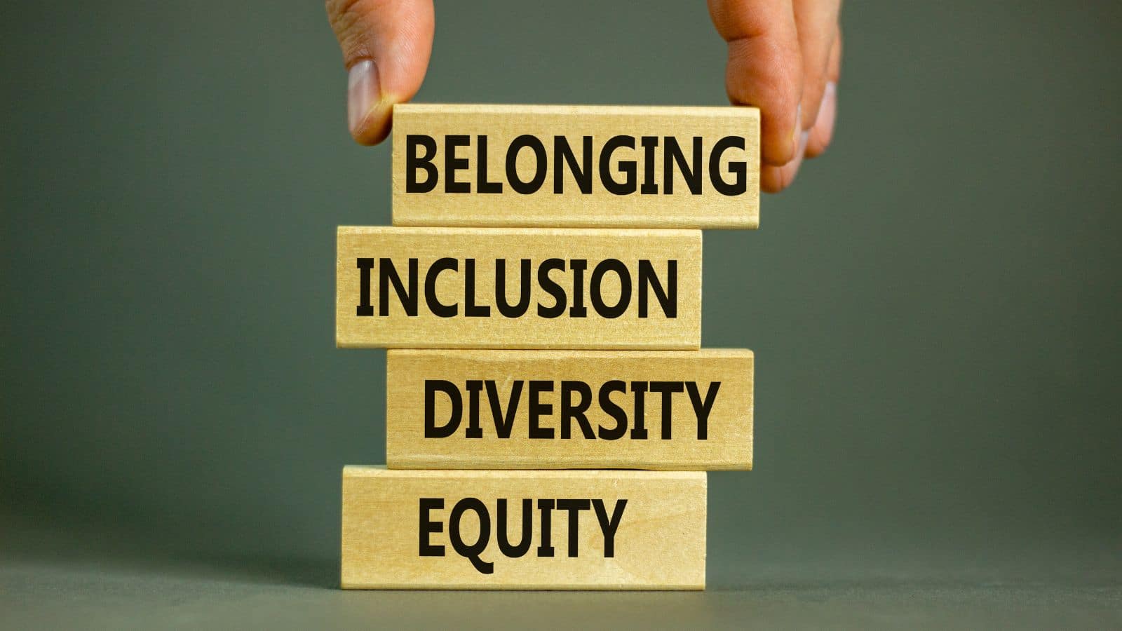 A hand placing a wooden building block onto a small stack that has the words 'Equity', 'Diversity', 'Inclusion' and 'Belonging'