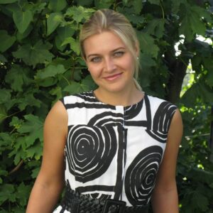 Head shot of smiling blonde woman, head tilted, against a green foliage